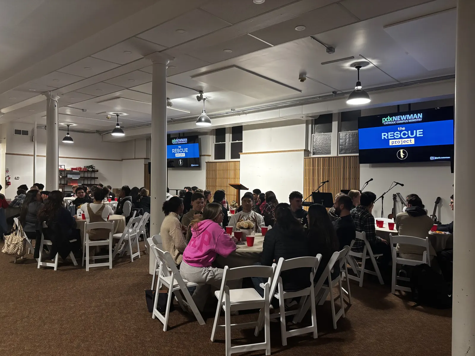 Students talking at round tables in the PDX Newman Center hall, with The Rescue Project title on the screens